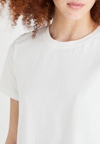 Hvid bomulds T-shirt med rund hals, korte ærmer, blød tekstur og minimal syning. Tæt på visning, der viser stof og pasform.