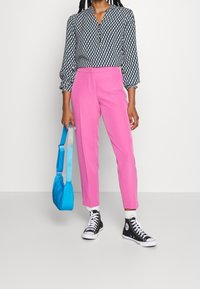 Blouse à motifs noir et blanc avec des manches trois-quarts, pantalon rose ajusté, baskets montantes noires et sac à épaule bleu.