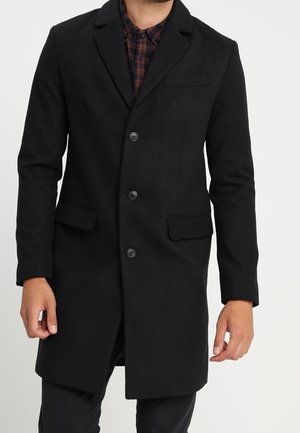 Homme portant un pardessus noir croisé à trois boutons par-dessus une chemise à carreaux rouge et noir.