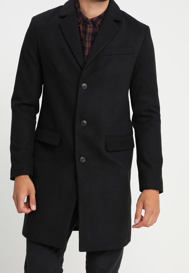 Homme portant un pardessus noir croisé à trois boutons par-dessus une chemise à carreaux rouge et noir.