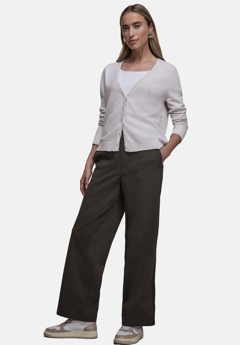 Cardigan beige chiaro con scollo a V e chiusura con bottoni, abbinato a pantaloni larghi scuri e sneakers bianche con dettagli beige.