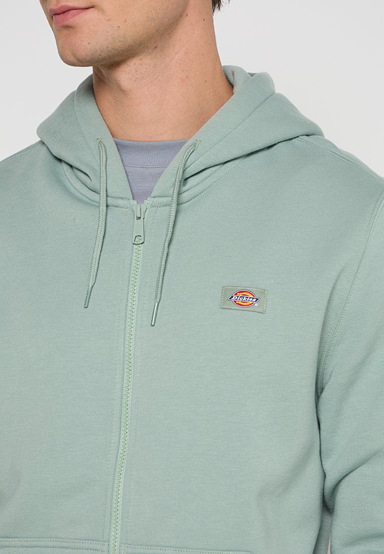 Sudadera con cremallera de color verde claro con capucha ajustable. Presenta un discreto parche de logo en el pecho y una textura suave de mezcla de algodón.