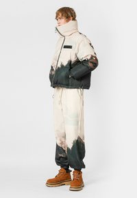 Chaqueta acolchada con estampado de montaña en beige y negro, combinada con pantalones a juego y botas marrones. Cuenta con cuello alto y cierre de cremallera.