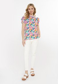 Farbige Bluse mit kurzen Ärmeln und Rüschenschultern, versehen mit einem tropischen Muster in Rosa-, Grün- und Orangetönen, kombiniert mit weißen Hosen und Sandalen.