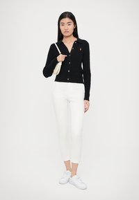 Černý žebrovaný kardigan s límečkem a zapínáním na knoflíky, doplněný o bílé přiléhavé kalhoty a bílé tenisky, držený s malou kabelkou.