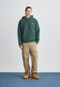 Sudadera verde con bolsillo frontal y logo, pantalones cargo beige con bolsillos laterales y botas marrones con cordones amarillos sobre fondo de alfombra azul.