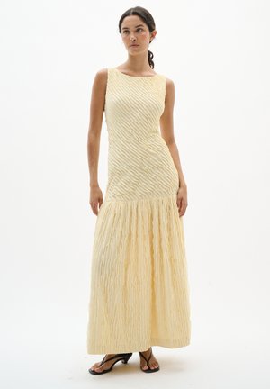 Nő áll egy ujjatlan, texturált halvány sárga maxi ruhában, amelynek testhezálló a felső része és bővebb az alja, fekete magassarkú szandált visel.