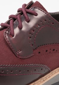 Burgundské oxfordské boty s kombinací lesklé kůže a semiše, doplněné brogue perforacemi a odpovídajícími kulatými tkaničkami.