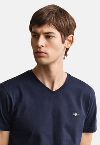 Jeune homme aux cheveux courts et bruns portant un T-shirt bleu marine à encolure en V avec un petit logo blanc et rouge sur la poitrine, regardant sur le côté.