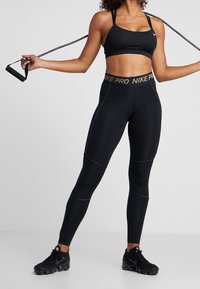 Kobieta w czarnym stanik sportowym, legginsach Nike Pro i butach, trzymająca skakankę za ramionami w pozie fitness.