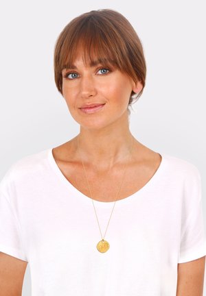Goldenes Anhänger-Halsband an einer zarten Kette, mit einem texturierten runden Anhänger. Getragen über einem weißen lässigen T-Shirt mit weitem Ausschnitt.