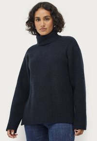 Donkerblauw gebreid coltrui met ribbeltextuur, verlaagde schouders en lange mouwen. De stof lijkt zacht en warm.