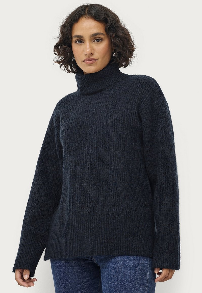 Donkerblauw gebreid coltrui met ribbeltextuur, verlaagde schouders en lange mouwen. De stof lijkt zacht en warm.