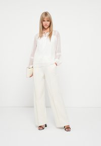 Blouse blanche avec des manches transparentes à pois, col à cordon. Associée à un pantalon large crême taille haute et des talons noirs et blancs à motifs.