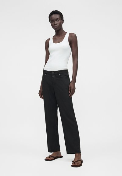 Femme debout portant un haut blanc sans manches côtelé, un pantalon large noir et des sandales noires contre un fond uni.