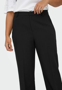 Pantaloni neri su misura con una texture liscia, dettagli delle pieghe e passanti per cintura. Si abbinano a una camicia bianca con accenti di perle.