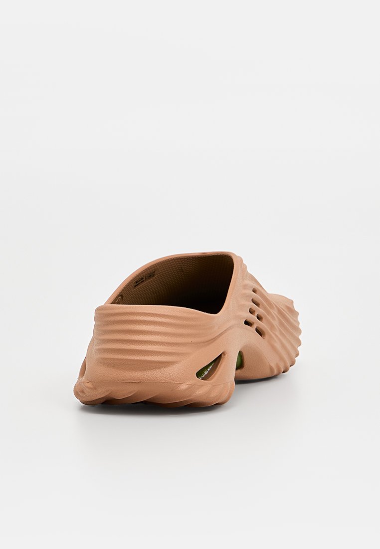 Scarpa slip-on marrone con superficie testurizzata e ondulata e fori di ventilazione sui lati. Suola piatta e design aperto sul retro.