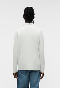 Μακρυμάνικη, ανοιχτό γκρι πόλο μπλούζα με υφή υφάσματος, γιακά και ευθεία ραφή, σε συνδυασμό με μπλε τζιν.