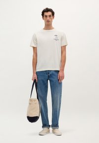 Creme kortærmet T-shirt med lille blå grafik på brystet, parret med lyst blå lige ben jeans, beige taske med marinebund og lyse sko.