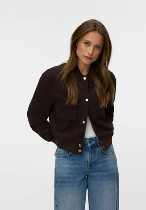 Kvinde med langt brunt hår iført en sort knappet jakke og blå jeans, stående foran en ensfarvet lys baggrund.