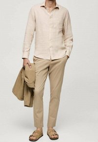 Camicia di lino beige chiaro con chiusura a bottoni e taschino, abbinata a pantaloni beige e sandali marroni, mentre tiene in mano una giacca coordinata color nocciola.