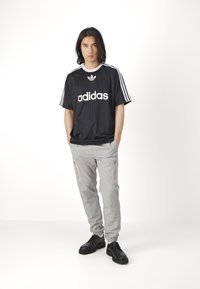 Fekete Adidas mez fehér logóval és csíkokkal, szürke, keskenyített nadrággal, amely cipzáras zsebekkel rendelkezik, és fekete cipőkkel párosítva.