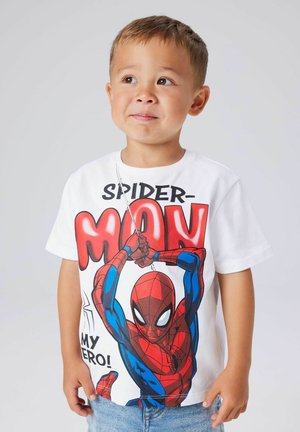 Niño pequeño con una camiseta blanca de Spider-Man que muestra una imagen de Spider-Man balanceándose en su telaraña y el texto "¡Mi héroe!" sobre un fondo liso.
