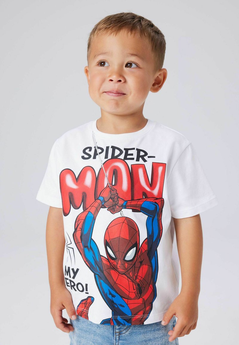 Νεαρός αγόρι που φοράει λευκό μπλουζάκι Spider-Man με γραφικό του Spider-Man να κουνιέται σε ιστό και κείμενο "Ο ήρωάς μου!" σε απλό φόντο.