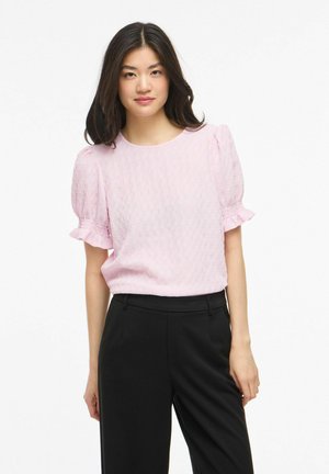 Młoda kobieta z długimi ciemnymi włosami, ubrana w teksturowaną jasnoróżową bluzkę z bufkami oraz czarne spodnie z wysokim stanem na białym tle.