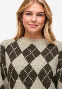 Pull en tricot beige clair avec motif argyle marron foncé. Présente un col rond et un matériau en laine texturée pour la chaleur.