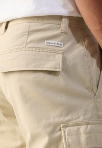 Shorts cargo beiges avec une texture lisse, dotés d'une poche rabat et d'une étiquette de marque "Marc O'Polo" cousue à l'arrière.