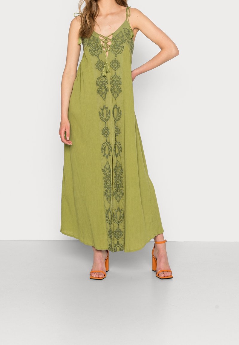 Femme portant une robe longue verte sans manches avec broderies florales et des sandales à talons hauts orange, debout devant un fond blanc.