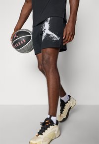 Sorte mesh basketballshorts med hvid grafisk print, parret med beige og sorte sneakers, og holder en mønstret basketball.