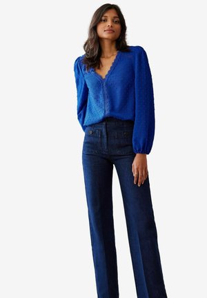 Helles blaues, strukturiertes Oberteil mit aufgeblähten Ärmeln; kombiniert mit dunklen Jeans mit geradem Bein und zwei Gesäßtaschen.