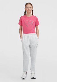 Jong meisje met gevlochten haar, gekleed in een roze t-shirt met logo, lichtgrijze sweats en witte sneakers, staande met handen in de zakken.