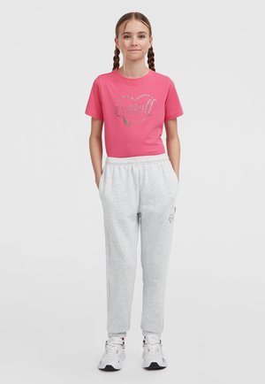 Junges Mädchen mit geflochtenem Haar, das ein pinkes T-Shirt mit Logo, hellgraue Jogginghosen und weiße Turnschuhe trägt. Sie steht mit den Händen in den Taschen.