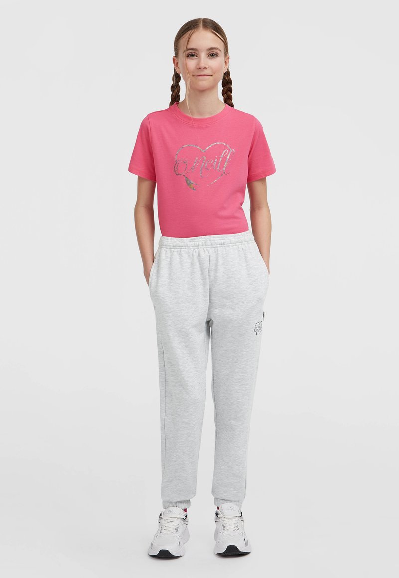 Jong meisje met gevlochten haar, gekleed in een roze t-shirt met logo, lichtgrijze sweats en witte sneakers, staande met handen in de zakken.