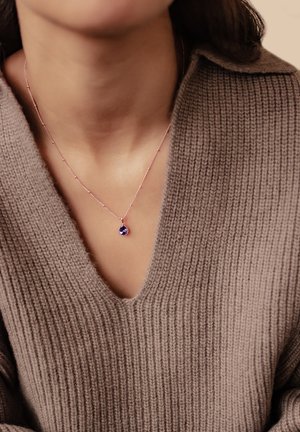 Roségoldene Halskette mit einem tropfenförmigen lila Edelsteinanhänger, getragen auf einem braunen, gerippten V-Ausschnittpullover.