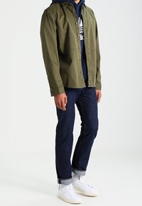 Person som bär en olivgrön skjorta med knappar över en marinblå huvtröja, mörkblå uppvikta jeans, vita strumpor och vita sneakers, stående på vit golv.