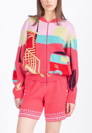 Vrouw draagt een kleurrijke trui met rits en een schilderachtig ontwerp, gecombineerd met bijpassende koraalkleurige korte broek met witte patroonaccenten.