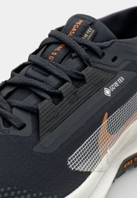 Sorte løpesko med teksturert stoff, GORE-TEX-detaljer, sorte snøringer, mesh-aksenter og en oransje logo. Nærbilde av funksjoner.