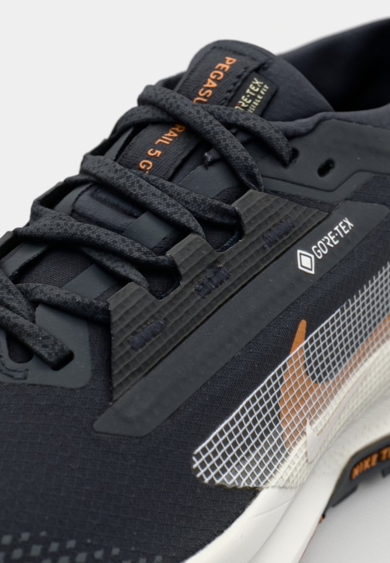 Zapatilla de running negra con tejido texturizado, detalles GORE-TEX, cordones negros, acentos de malla y un logo naranja. Vista cercana de las características.