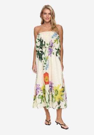 Młoda kobieta w kremowej, bezramiączkowej, zwiewnej sukience z dużym, kolorowym kwiatowym wzorem, w czarnych sandałach na obcasie, stoi na tle białego tła.