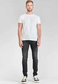 T-shirt bianca a maniche corte abbinata a jeans slim-fit scuri. Le sneakers sono di colore bianco e nero con design a taglio basso. Sfondo uniforme.