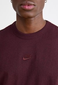 Marooni puuvillane T-särk, millel on ümar kaelus ja rinnal väike, samas toonis tikandina Nike logo. Sile tekstuur.