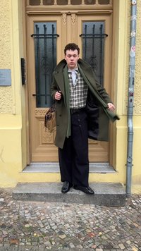 Soprabito verde, camicia a quadri, pantaloni scuri, scarpe nere. Tiene una borsa marrone. In piedi accanto a un percorso lastricato di pietra e una porta di colore giallo chiaro.