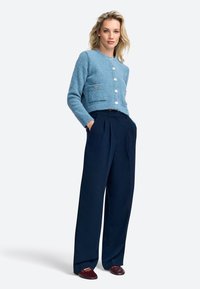 Cardigan texturé bleu clair avec un col rond, quatre boutons et deux poches avant, associé à un pantalon plissé marine et des mocassins bordeaux.