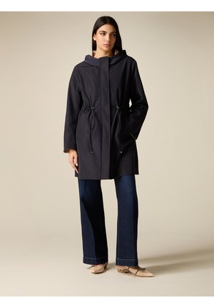 Nainen, jolla on yllään musta huppari, jonka vyötäröllä on kiristysnauha, leveälahkeiset tummansiniset farkut ja beiget terävät kengät, seisoo yksinkertaisen taustan edessä.