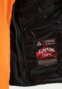 Μαύρη εσωτερική επένδυση σε μια μονωμένη πορτοκαλί ζακέτα με ετικέτα που την περιγράφει ως Nike ACG μονωμένο μπουφάν τρεξίματος με Therma-Fit AeroLoft.