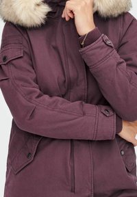 Bordowa kurtka parka z futrzanym kapturem, zapinana na zamek i guziki, z bocznymi kieszeniami i teksturowaną powierzchnią.